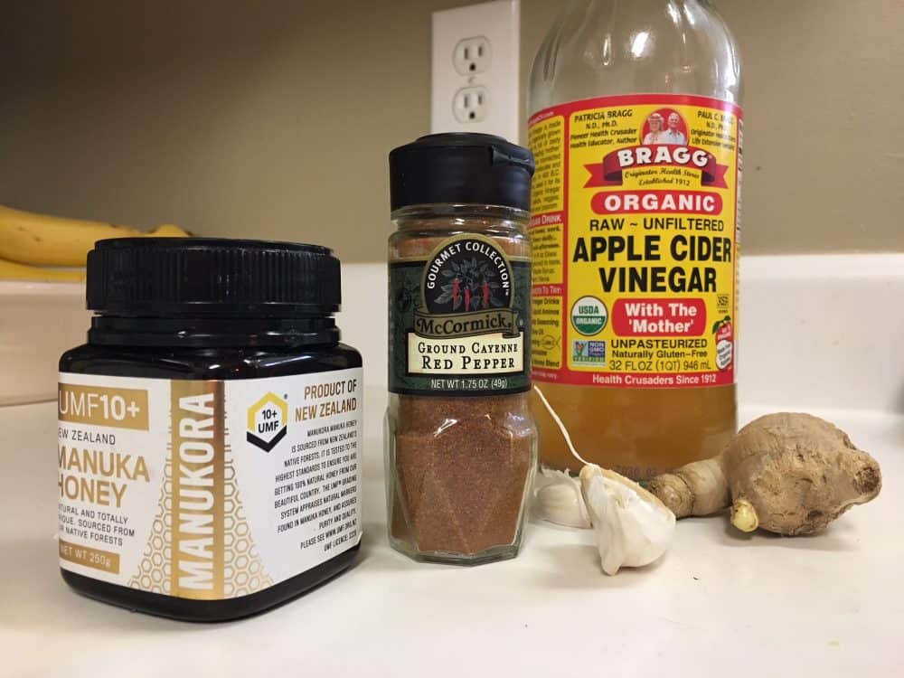 Ingredients for how to cure a cold with super tonic: manuka honey, cayenne pepper, apple cider vinegar bottles with ginger and garlic on a kitchen countertop.