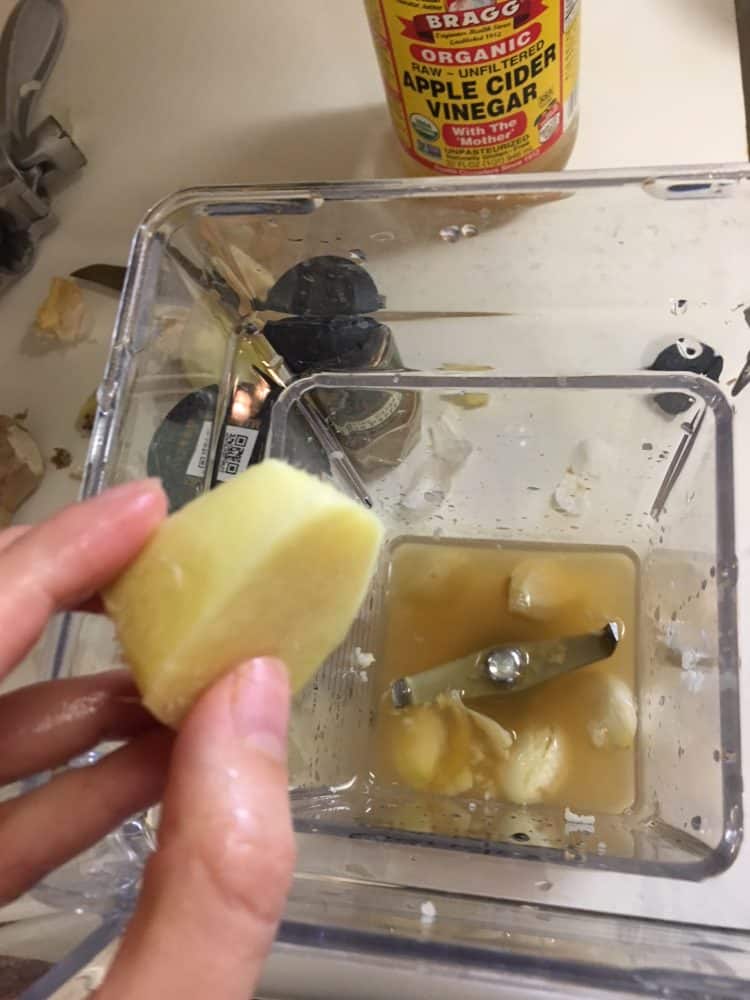 Adding ginger root to a blender filled with apple cider vinegar and fresh garlic.