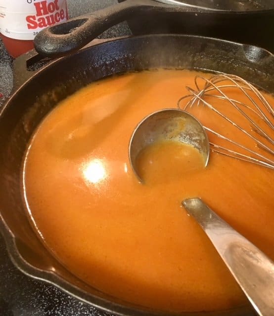 Chicken wing sauce in a cast iron skillet, with a whisk and ladle sitting inside the pan.