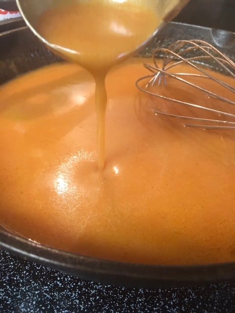 Chicken wing sauce in a cast iron skillet, being poured slowly into the pan with a ladle.