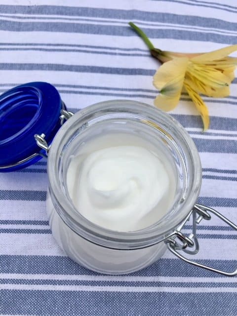 All natural lotion in a small jar on a striped tea towel beside a yellow blossom.