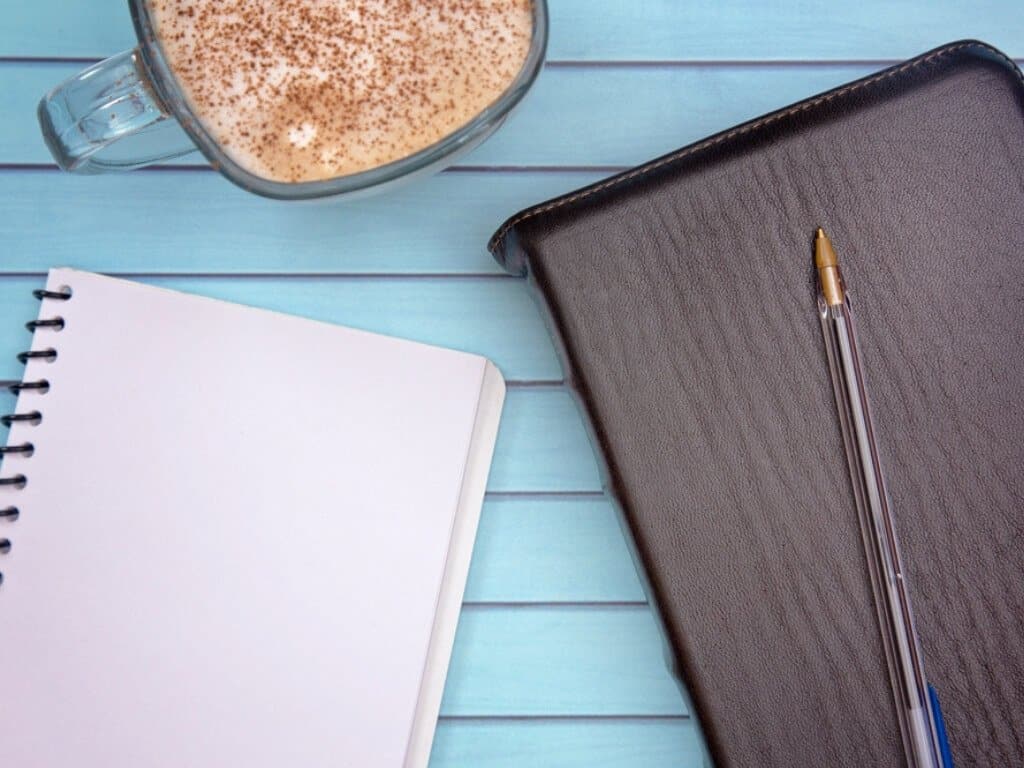 Prayer journaling notebook, pen, bible, and cappuccino sitting on a blue wooden table.