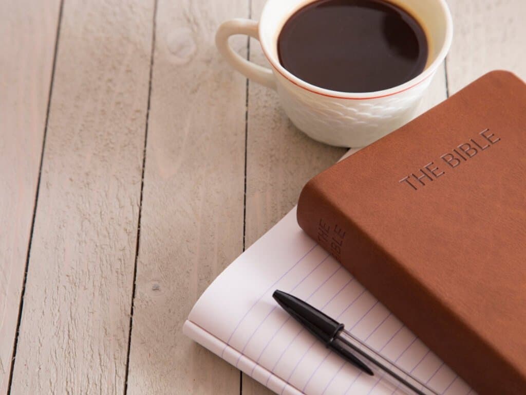 Prayer journal, pen, bible and cup of coffee sitting on a rustic table.