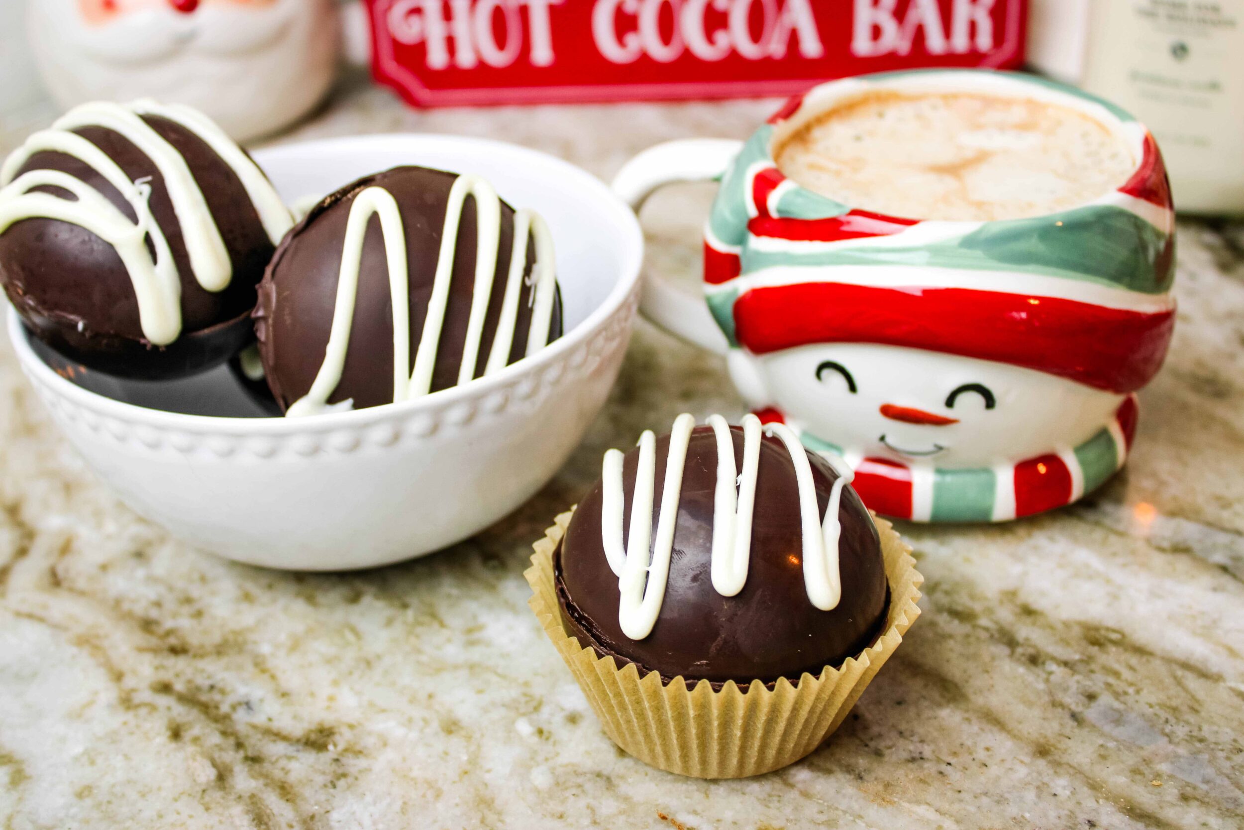 Hot cocoa bomb sitting on a countertop next to more cocoa bombs and a cup of hot cocoa.